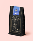 A black and gold coffee bag labeled “Magic Hour Time for Infinity,” adorned with an hourglass illustration, blue seal, and gold star patterns, rests against a pale pink background—capturing the enchanting appeal of the Moringa Moon Herbal Tea Refill Pouch.