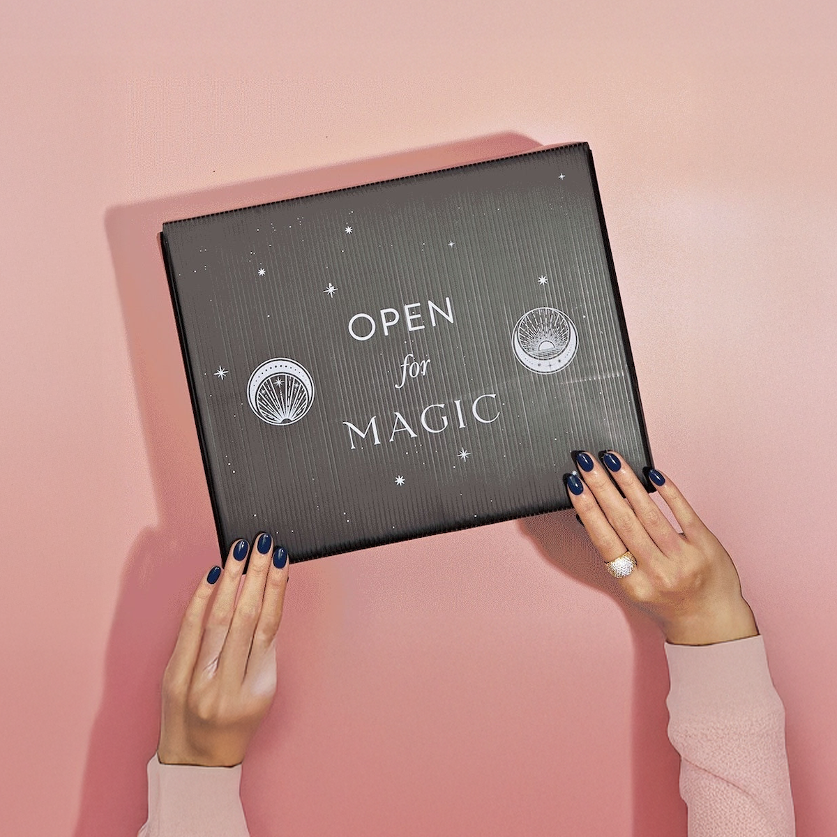 A person with dark blue nails and a ring holds the Labor Day Mystery Box against a pink background. The black box says "OPEN for MAGIC" in white, with star and shell designs, hinting at hand-selected teas inside.