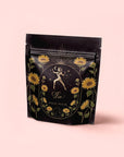 A black resealable bag with gold and yellow floral accents depicts a woman holding a star, labeled "Leo" and "Magic Hour," containing Leo: Queen of Courage Tea of the Sun (Yunnan Black Tea), set against a pink background.