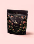 A black resealable pouch with floral designs, gold accents, and an illustration of a woman and scorpion beneath a starry sky features the label “Scorpio Tea for Sensual Brilliance.” Set against pastel pink—perfect for lovers of sensual tea.