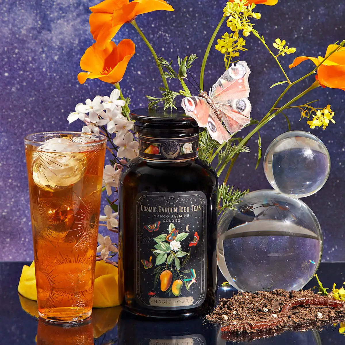 A tall glass of iced tea with ice sits beside a dark bottle labeled "Cosmic Garden Iced Teas: Mango Jasmine." Bright orange flowers, white blossoms, a pink butterfly decoration, a small glass orb, and soil with seeds complete the vibrant tabletop scene.