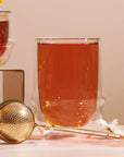 A glass of tea sits on a table beside a Luxe Loose Leaf Strainer and a small white flower, with another cup partially visible in the background.