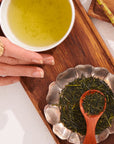 A manicured hand rests by a cup of Japanese green tea and a metal dish of Flower Fragrance Iccha Kariban Green Tea leaves on a wooden board. A wooden spoon sits atop the vibrant loose leaves, highlighting this premium Kagoshima tea.