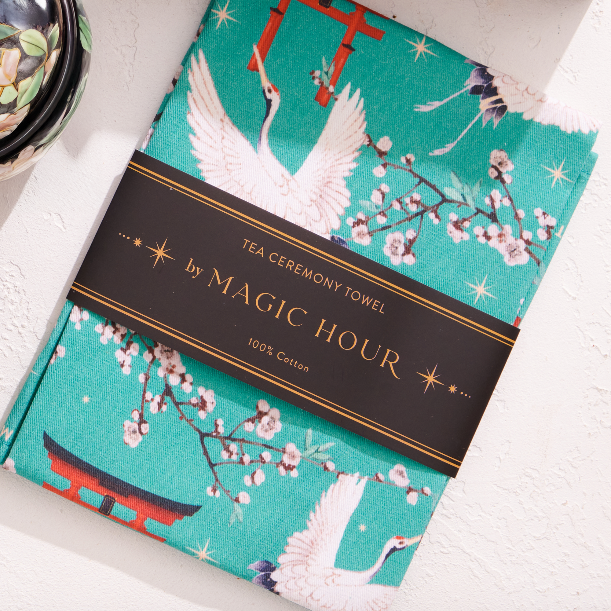 The Kyoto Tea Towel, inspired by Japan, showcases teal designs of white cranes, cherry blossoms, and red torii gates. Named the "Tea Ceremony Towel by Magic Hour," it adds a Japanese flair to your home and is made from 100% cotton.