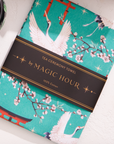 The Kyoto Tea Towel, inspired by Japan, showcases teal designs of white cranes, cherry blossoms, and red torii gates. Named the "Tea Ceremony Towel by Magic Hour," it adds a Japanese flair to your home and is made from 100% cotton.