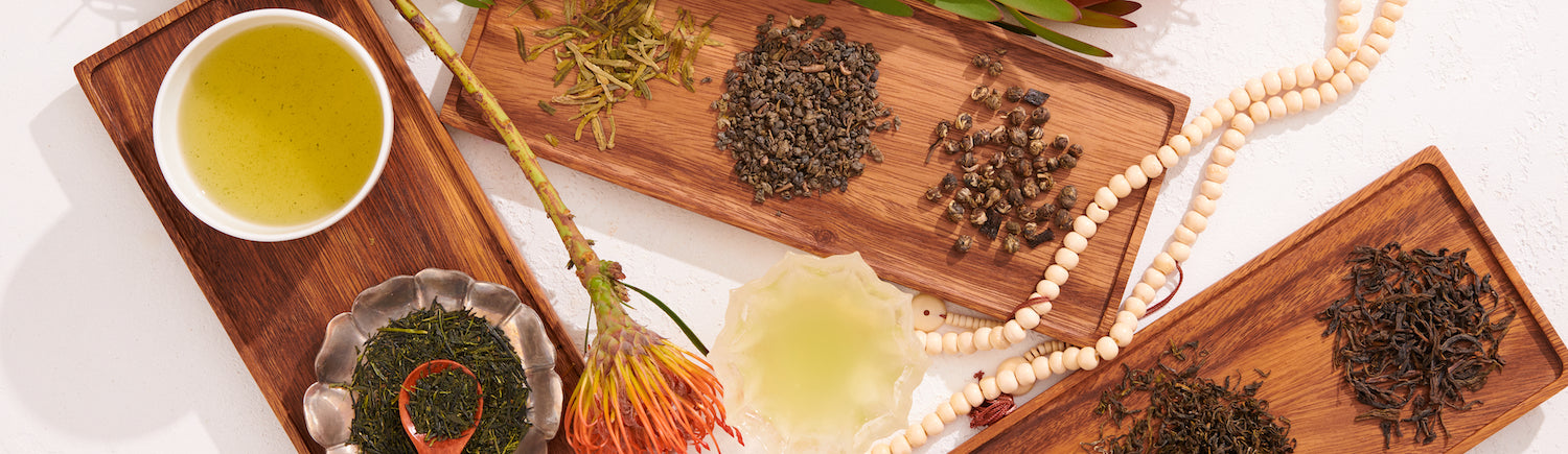 Wooden trays display various types of loose tea leaves and herbs, including a steaming cup of green tea, a wooden spoon, a flower, and a string of white beads, all arranged artistically on a light-colored surface.
