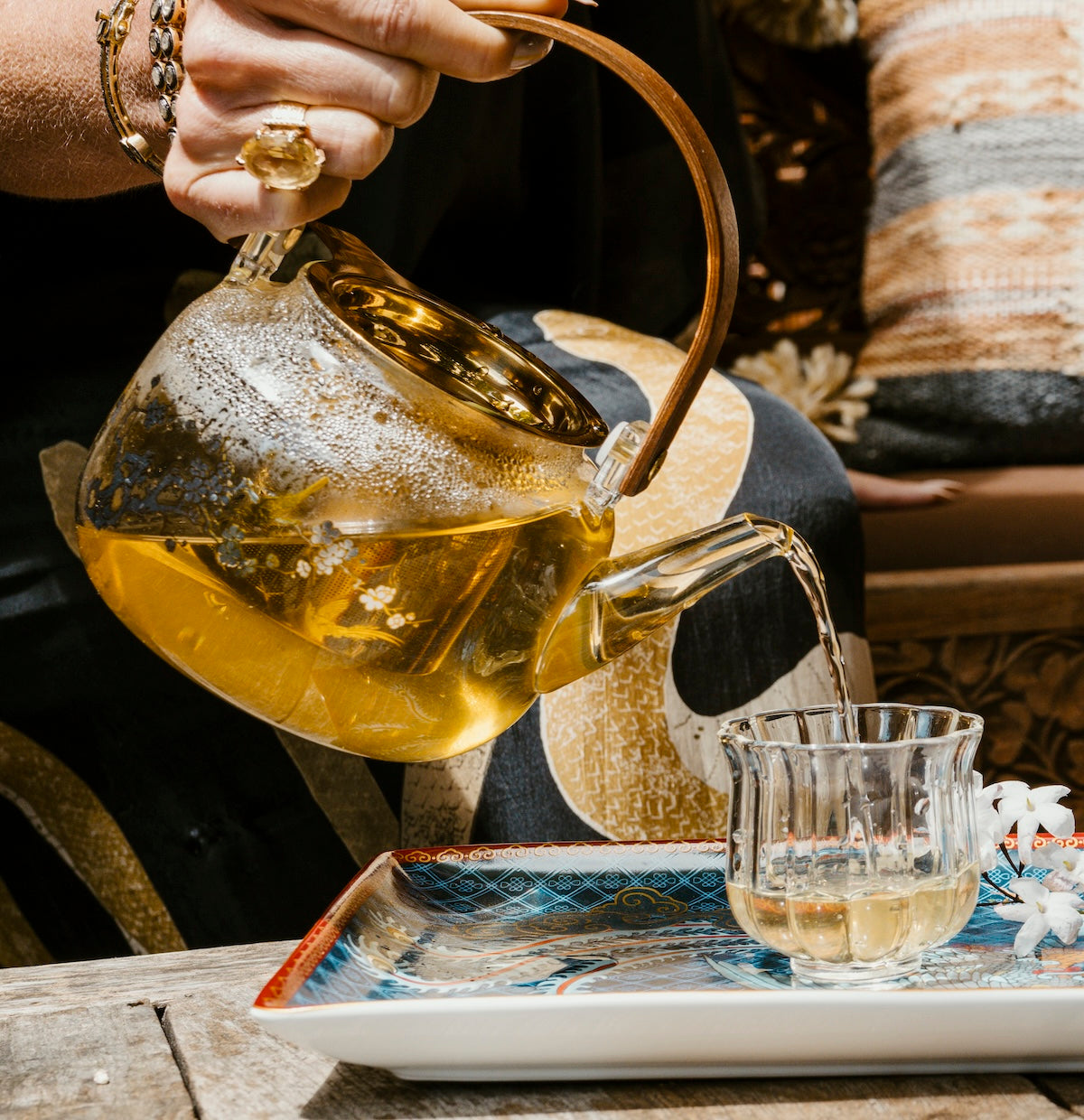A person pours Dragon Yuzu Oolong tea from a glass teapot into a clear glass on a wooden table. The colorful tray beneath the glass adds visual interest, as the person, adorned in a flowy garment with an attention-grabbing ring and bracelet, completes the ritual. The background is filled with comfortable pillows and artistic touches, subtly evoking the calming aroma of Japanese Yuzu Essential Oil from Magic Hour’s Nuwa Tea for Intuition line.