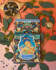 A box of Magic Hour's Nuwa Tea for Intuition: Dragon Yuzu Oolong Case Packs, adorned with intricate artwork of a meditative figure, is surrounded by green leaves, small purple flowers, a tea infuser, and loose Taiwanese Tung Ting-Dragon Oolong on a gradient orange-to-pink background.