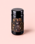A black cylindrical jar adorned with a striking label reading “Gemstone Wellness Tea Black Onyx” rests on a soft pink surface, inviting you to explore its caffeine-free elixir—a restorative blend expertly crafted with Chaga, Maca, and other nourishing ingredients for energy, immunity, and good fortune.