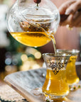A hand pours yellow herbal tea—likely from the Oolong Tea Sampler Box—into ornate glass cups on a metal tray, with small white flowers scattered around.