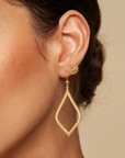 Close-up of a woman's ear adorned with 18kt gold plate earrings. One earring is a small sun-shaped stud, and the other is a larger, diamond-shaped drop earring featuring an elegant, intricate design from Magic Hour's Open to Beauty Green Onyx Earrings. A hint of green onyx gleams from the background's neutral tone.