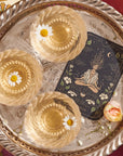 A decorative silver tray holds three translucent jelly desserts, each topped with a white daisy. Beside the desserts, there is a tarot card featuring a meditative figure surrounded by nature. A Magic Hour's "The Oracle: Lemon Lavender Tea for Supporting the Gut-Brain Axis" caffeine-free tea bag rests near the edge of the tray alongside a pink and white flower.