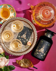 An elegant setup featuring a decorative tray with three glasses of champagne, a black bottle with an intricate label, Magic Hour's The Oracle: Lemon Lavender Tea for Supporting the Gut-Brain Axis in a clear teapot, a floral teacup, and a golden spoon with loose tea leaves, all arranged on a pink surface alongside Adaptogenic roots and white roses.