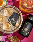 An elegant setup featuring a decorative tray with three glasses of champagne, a black bottle with an intricate label, Magic Hour's The Oracle: Lemon Lavender Tea for Supporting the Gut-Brain Axis in a clear teapot, a floral teacup, and a golden spoon with loose tea leaves, all arranged on a pink surface alongside Adaptogenic roots and white roses.