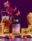 A bottle labeled "Pitta Herbal" is displayed with a teapot, teacup, honey jar, cinnamon sticks, flowers, a golden Buddha head statue, and brass utensils against a purple background.