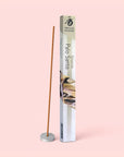 A Natural Incense : Palo Santo stick stands upright in a small holder beside its rectangular packaging, set against a pale pink background.