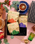 Peace & Joy Sampler Set featuring three tea pouches in a black box surrounded by a cup of tea, loose tea, a tart, a tea strainer, and two decorative glasses.