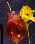 A glass of iced tea with the Sips Tea Straw & Strainer Bombilla, garnished with a bright yellow flower, sits against a starry night background. A bottle labeled “Spiced Teas” is partially visible on the left, hinting at the rich flavors of loose tea within.