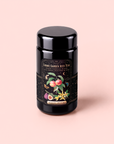 A black tea canister labeled “Cosmic Garden Iced Teas, Peach Vanilla Black Iced Tea with Figs,” adorned with colorful fruit and flower illustrations, sits on a pale pink surface—perfect for those seeking a touch of Peaches & Cream tea delight or a flavorful digestive wellness tea.