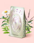 The "Pearl Tea of Beauty, Clarity, and a Sense of Peace" package, adorned with pastel colors, purple stars, and an ornate label, stands in front of illustrated chamomile, mint, and Tulsi plants against a light pink background.