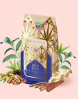 A decorative bag of Magic Hour Pecan Pie Chai herbal tea with illustrated leaves, surrounded by chai spices, cinnamon sticks, ginger root, rooibos, and other herbs on a pale pink background.