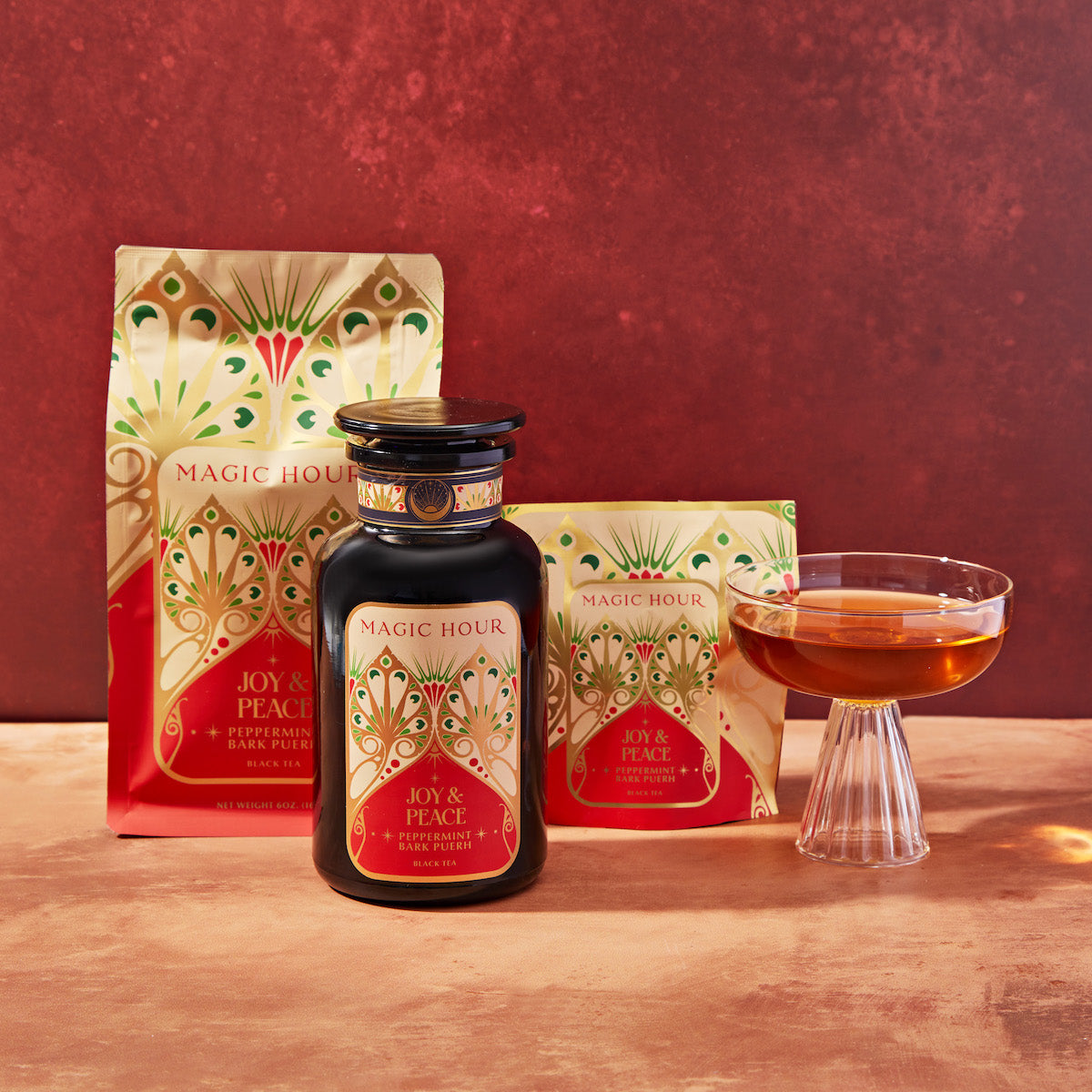 A product display includes a decorated red and gold tea bag, a black tea jar, and a cup of brewed tea. The packaging is adorned with intricate designs and labeled "Magic Hour Joy & Peace Peppermint Bark Pu-erh". The background is a warm, rich reddish-brown.