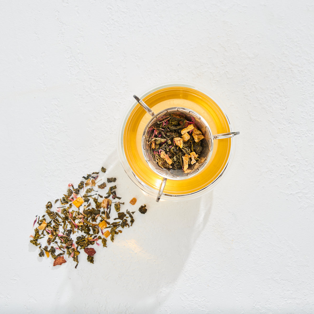 Top view of a glass teacup with yellow tea, a tea infuser inside holding loose tea leaves. Some of the loose tea leaves are spread out on the white surface next to the cup. The image has bright lighting and a minimalist aesthetic.
