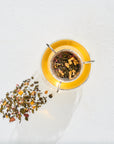 Top view of a glass teacup with yellow tea, a tea infuser inside holding loose tea leaves. Some of the loose tea leaves are spread out on the white surface next to the cup. The image has bright lighting and a minimalist aesthetic.