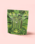 A green, gemstone-patterned pouch labeled "Peridot: Green Guayusa Oolong Mango-Peach Tea" stands upright against a light pink background, featuring a faceted gemstone design and highlighting its refreshing mango-peach flavors.