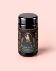 A black jar labeled “Magic Hour Persephone’s Potion: Pomegranate & Jasmine Green Tea,” featuring an illustration of a woman among flowers, set against a soft pink background—an enchanting blend inspired by the nourishing tradition of herbal tea for natural immunity support.