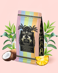 A coffee bag with a vibrant rainbow design, palm trees, and a black label that reads "Piña Colada: Tropical Green Mocktail Tea for Sultry Moods & Radiant Skin" is surrounded by coconut, pineapple, and lush green leaves—evoking the tropical notes of this pineapple coconut tea—set against a pale pink background.