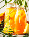 Two glass jars of tropical pineapple juice, adorned with pineapple slices and twine, sit among pineapple pieces and palm leaves, capturing a refreshing summer vibe inspired by Pineapple Oolong.