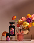 A vibrant still life composition features a golden vase with colorful flowers, a dark bottle and jar adorned with matching floral labels, and a glass decanter filled with pink liquid. The setup, which includes the immune-supporting *Pink Moment Pixie - Hibiscus Tangerine Dream Herbal Tea* by *Magic Hour*, is arranged on a beige background with golden platforms.