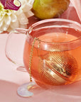 A glass mug of hot tea with a gold Opalite Moon Tea Strainer and a crescent moon charm sits on a pink surface, accompanied by a white flower, an orchid, and a pear in the background. Steam curls from the tea, enhancing the soothing ambiance of a spiritual tea ritual.