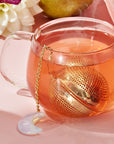 A clear glass teacup filled with reddish tea sits on a pink surface, paired with The Best of Fall Box. Inside, a gold mesh tea infuser attached to a chain with a crescent moon charm steams slightly, signaling hot tea. A white flower and part of a pear are visible in the background.
