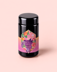 Introducing Pink Moment Pixie - Hibiscus Tangerine Dream Herbal Tea, a beautifully packaged black tea canister adorned with a colorful label of fruits and flowers on a light pink background. This vibrant, caffeine-free blend features tangerine hibiscus tea complemented by elderberry and rosehip for a delightful herbal tea experience.