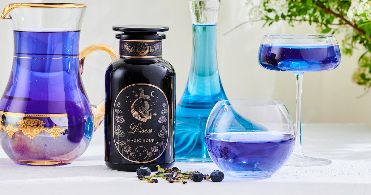 A collection of items including a bottle of "Pisces Magic Hour" tea, various glass containers filled with blue liquid, and some scattered tea leaves and blueberries, all arranged on a light surface with a floral background.