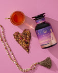 A bottle of Pitta Herbal supplements, a cup of tea, a heart-shaped pile of dried herbs, and a beaded tassel necklace are arranged on a pink background.