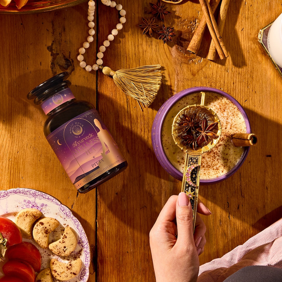 A hand holds a gold tea strainer with star anise over a spiced drink in a purple cup; next to it are Pitta Herbal supplements, cookies, spices, and beads on a wooden table.