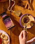 A hand holds a gold tea strainer with star anise over a spiced drink in a purple cup; next to it are Pitta Herbal supplements, cookies, spices, and beads on a wooden table.