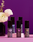 Five bottles of Planetary Perfume Oil, each with a celestial label, are displayed on pink next to a dark vase holding white flowers, all set against a purple backdrop—capturing cosmic elegance and aromatic allure.