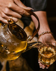 Close-up of a person pouring April 2024 Harvest - Grand Cru First Flush Organic "Spring Wonder" Samabeong Estate Darjeeling STGFOP1 from Magic Hour from a clear glass teapot into a small glass teacup. The teapot contains light-colored, organic tea from Samabeong Tea Garden, and steam can be seen rising. The person is wearing beaded bracelets and a ring on their finger.