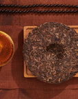 An Organic Puerh Tea Cake - 200g, renowned for its fermented intricacy, rests on a wooden tray next to a cup of amber-hued tea. Above them, a strand of dark beads stretches horizontally against a brown textured fabric backdrop.
