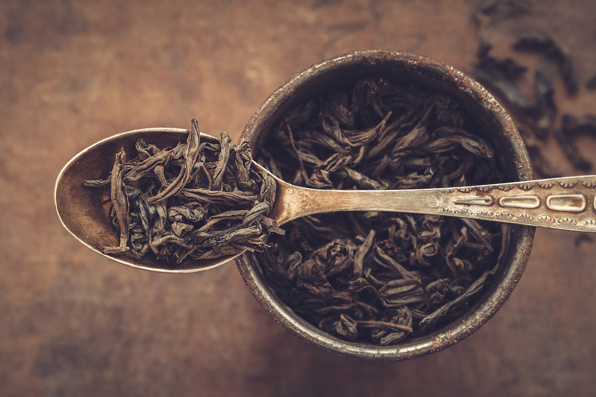 A close-up shot of loose-leaf tea in an antique metal spoon resting on the edge of a rustic metal cup filled with more loose-leaf tea. The background is a textured, earthy surface with some scattered tea leaves.