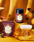 Pumpkin Spice Matcha jars and a drink are displayed on gold fabric surrounded by autumn botanicals, vibrant flowers, and pastries, creating a cozy fall atmosphere.
