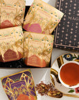 A stylishly arranged photo featuring a box of "Pumpkin Spice Abundance Sampler Bundle" tea packets in festive, decorative packaging. Next to the box is a teacup filled with tea, adorned with floral designs. Some tea leaves are scattered by the cup, adding a charming touch reminiscent of autumn's warmth and imaginative flavors.