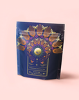 A navy blue tea pouch with vibrant floral and bird designs, gold accents, and a pale pink label that reads "Queen of the Rainforest: Cacao-Berry Tea," featuring an energizing rainforest blend.
