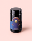 A black tea canister with a colorful sun and moon motif on a blue label reading “Queen of the Rainforest : Cacao-Berry Tea” sits against a pale pink background, perfectly capturing the vibrant rainforest energy found in every sip of this unique cacao-berry infused Guayusa tea.
