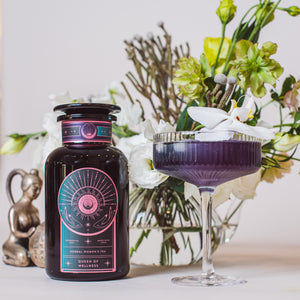 A dark glass bottle labeled "Queen of Wellness: Women's Hormone Balancing Tea for PMS, Healthy Cycles & Menopause" stands beside a purple drink with a white flower garnish, surrounded by flowers and a decorative statue.