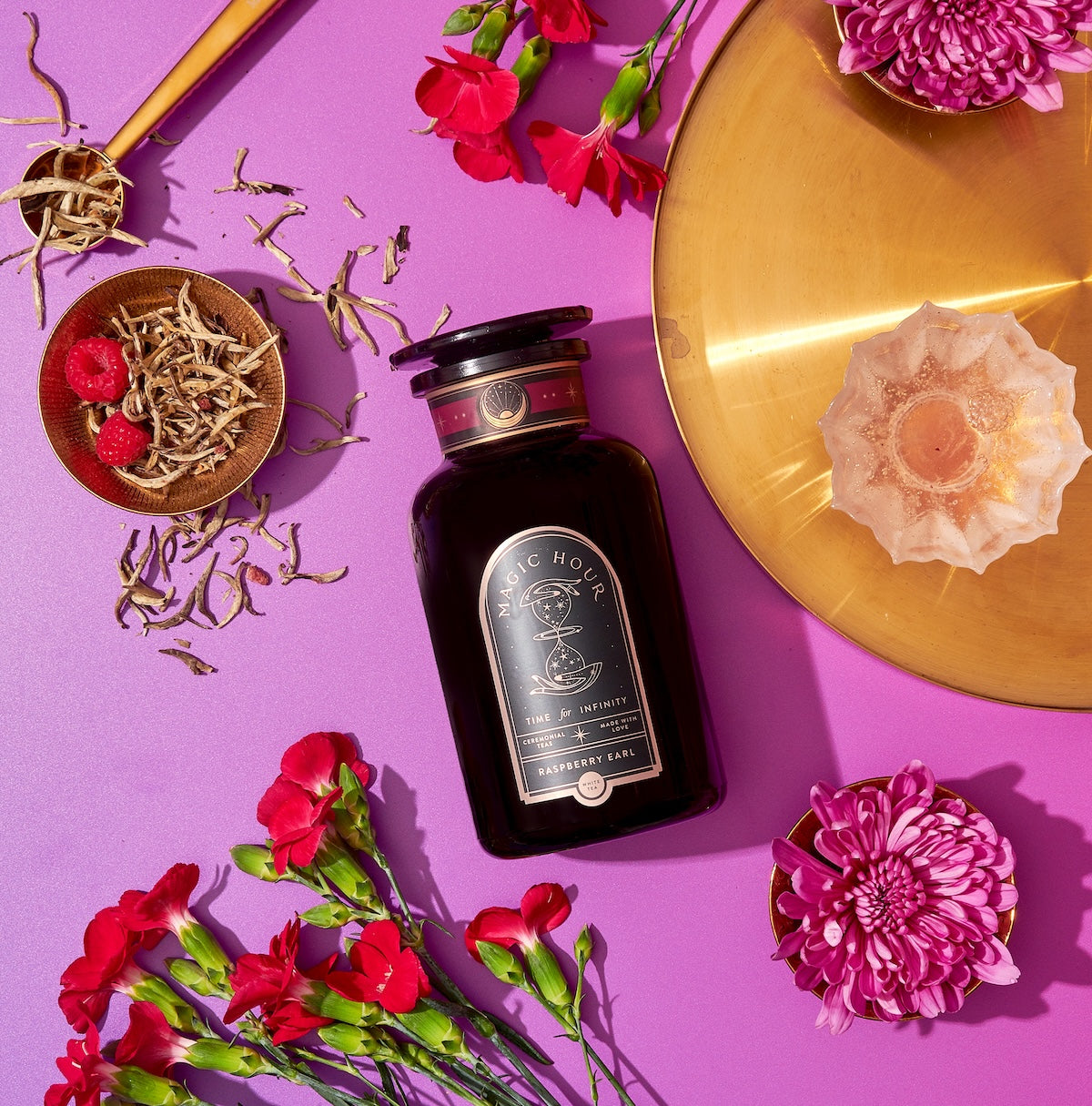 A black bottle labeled "Raspberry Earl White Tea" lies on a vibrant purple background, accompanied by a gold plate, scattered white tea leaves rich in antioxidants, red flowers, a small dish of raspberries, and a pink-lit candle in a rose-shaped holder.